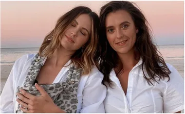 Two women in white shirts stand on a beach at sunset, one resting her head on the other's shoulder.