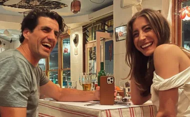 A man and woman laughing at a restaurant table, with drinks and a smartphone visible.