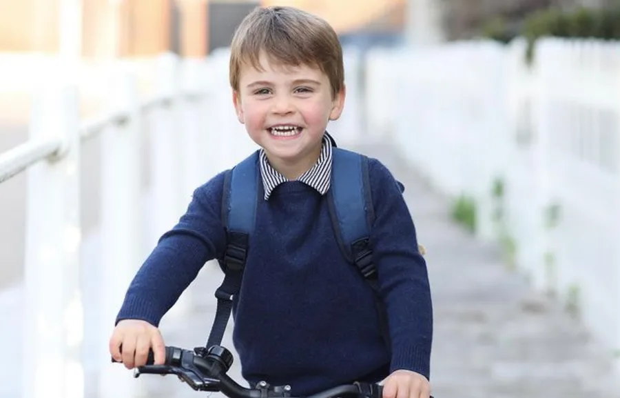 A young boy in a blue sweater joyfully rides a bicycle with a backpack on, along a path lined with white rails.