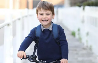 A young boy in a blue sweater joyfully rides a bicycle with a backpack on, along a path lined with white rails.