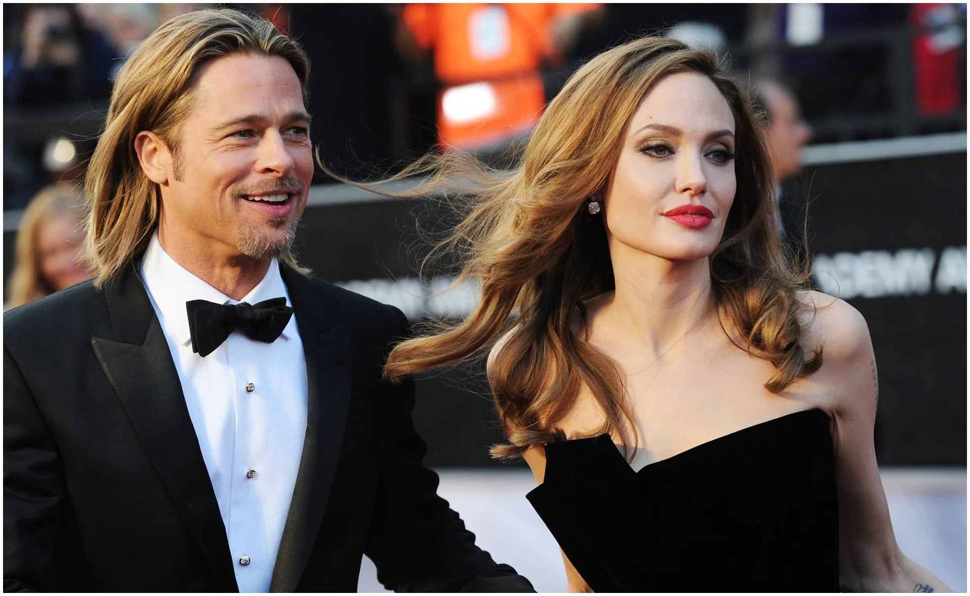 A man in a tuxedo and a woman in a strapless dress attend a formal event, with people in the background.
