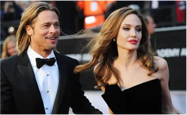 A man in a tuxedo and a woman in a strapless dress attend a formal event, with people in the background.