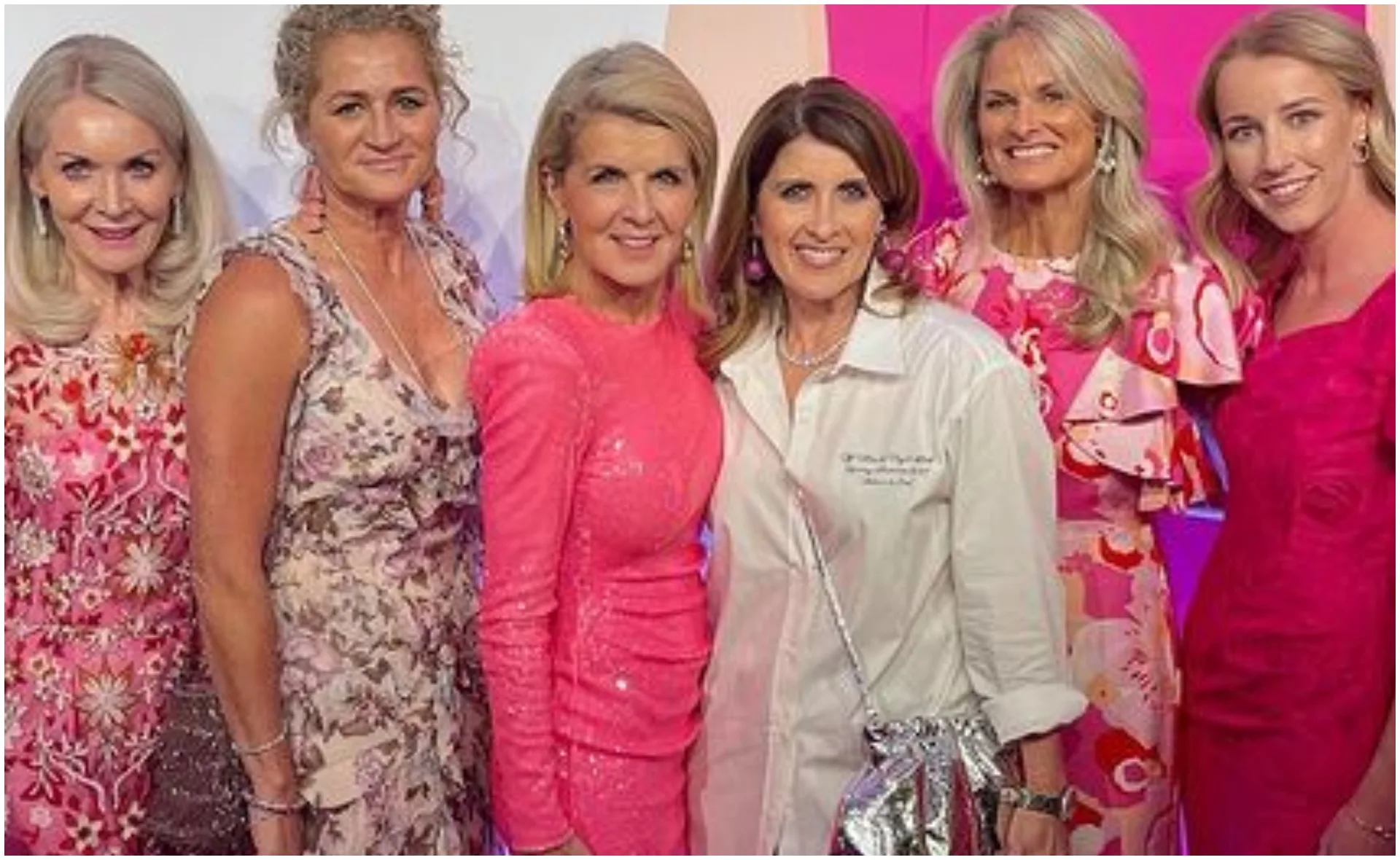 Group of six women smiling in colorful, elegant dresses at an event.