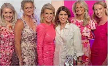 Group of six women smiling in colorful, elegant dresses at an event.