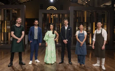Six people standing in a kitchen studio, with some wearing aprons and others dressed in smart-casual attire.