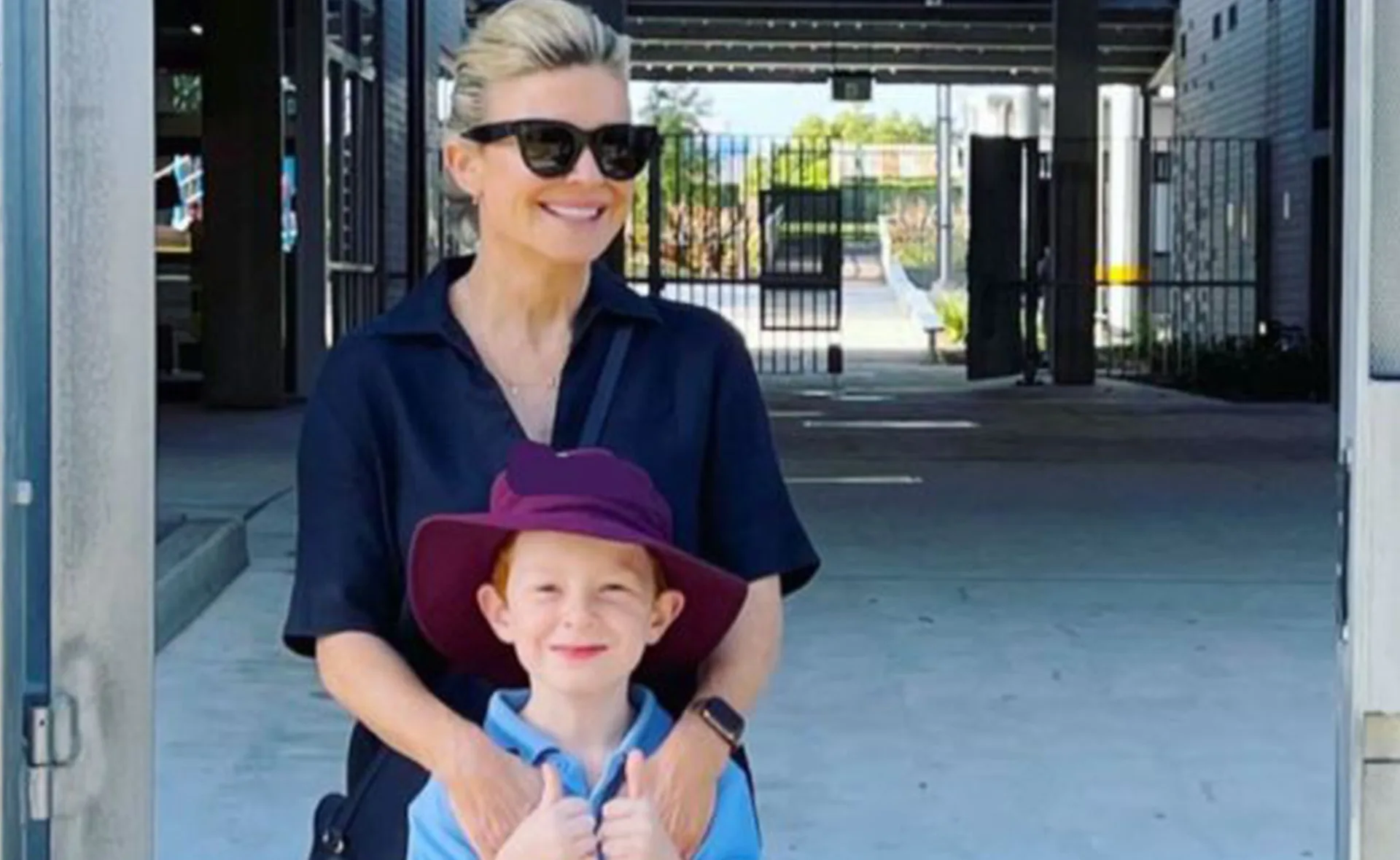 A woman in sunglasses with a young boy wearing a hat, standing in a courtyard.