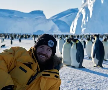 Man in yellow jacket smiling with penguins in snowy Antarctic landscape.