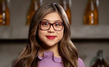 A woman with glasses and long hair, wearing a purple top, smiles in a well-lit room.