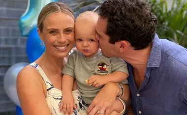 A family smiles with a child wearing an "Australia" patch shirt, as a man kisses the child's cheek, with balloons in background.