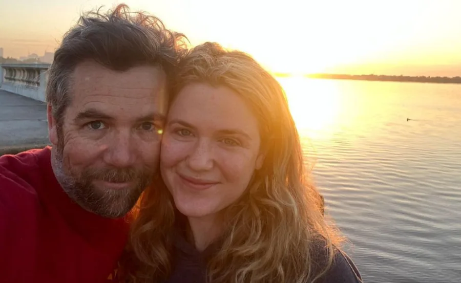 A couple smiling at the camera with a sunset over a lake in the background.