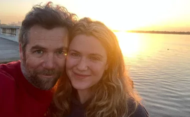 A couple smiling at the camera with a sunset over a lake in the background.