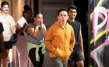 Group of people, some with surprised expressions, walk indoors; focus on person in orange hoodie.