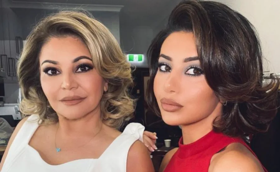 Two women with styled hair and makeup pose closely indoors, one in white and the other in red.