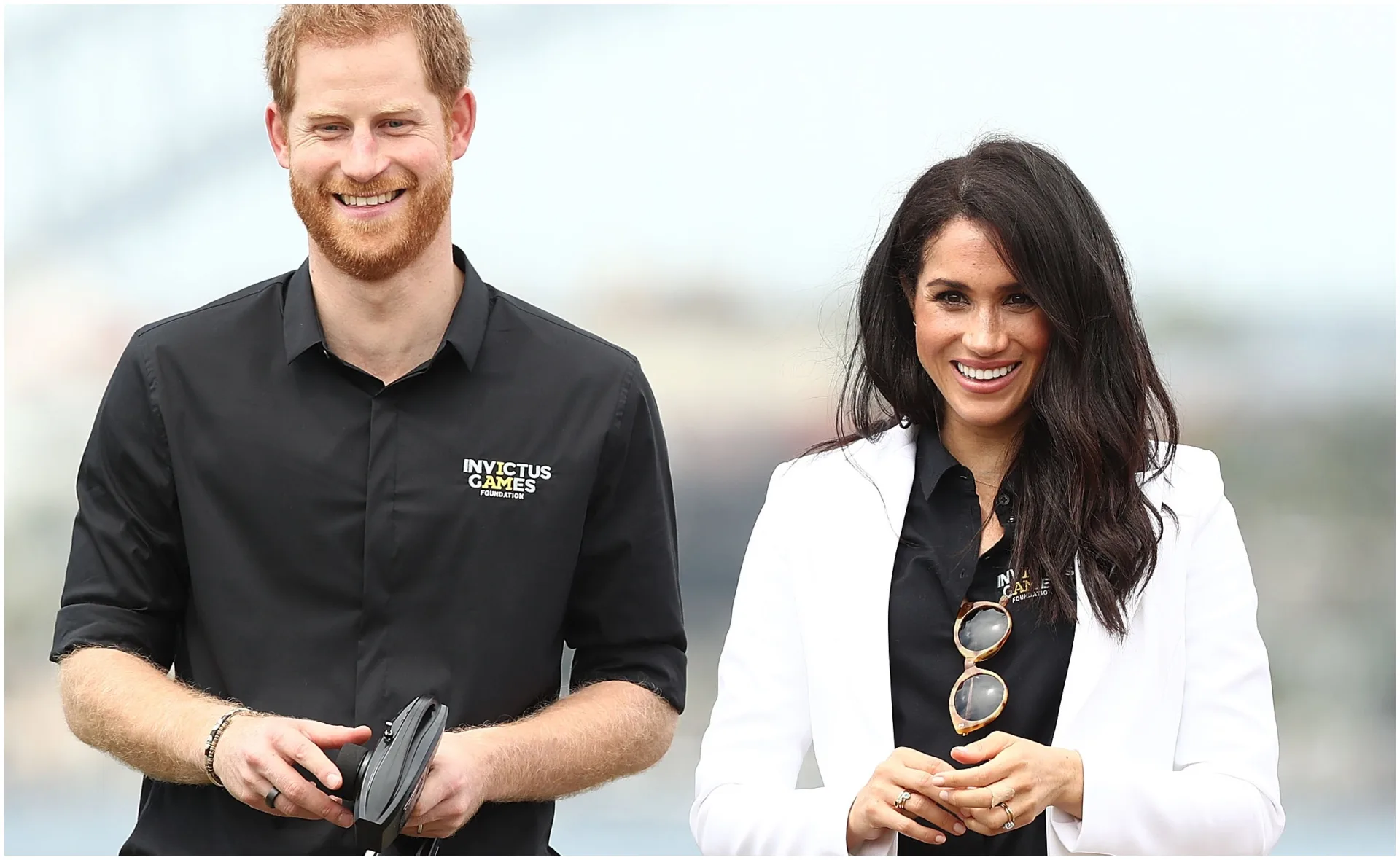 Two people smiling, one in a black shirt with "Invictus Games" logo, and the other in a white blazer holding sunglasses.