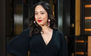 Woman with long wavy hair, wearing bold red earrings and a black outfit, in a softly lit interior setting.