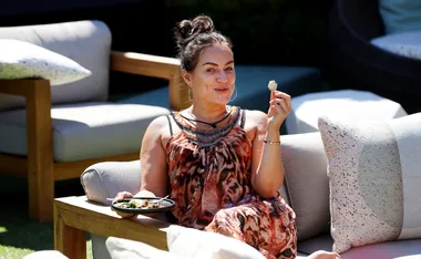 A woman sits on an outdoor couch enjoying a snack, wearing a patterned dress in a relaxed sunny setting.