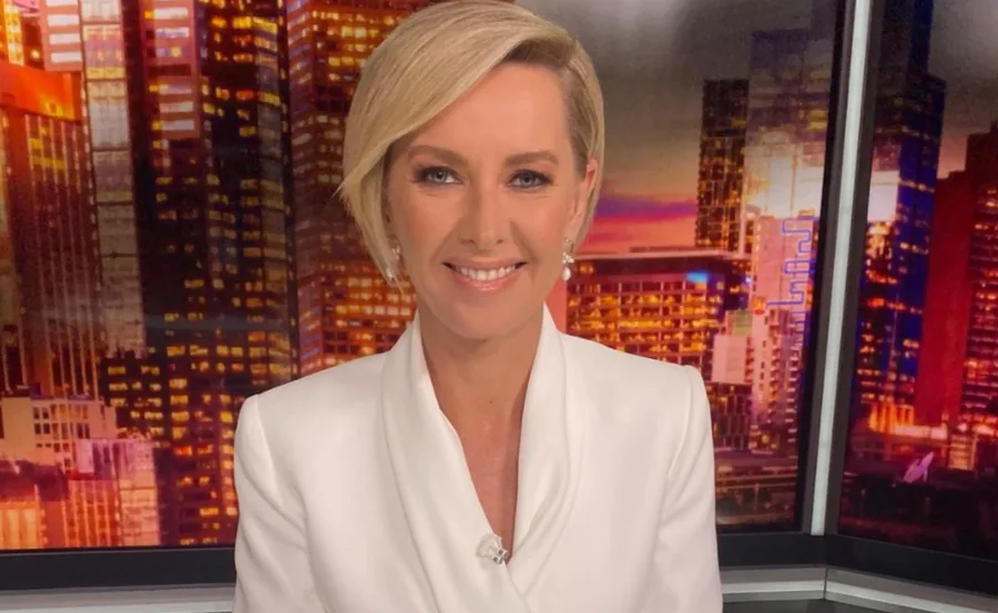 A person with short blonde hair in a white suit, smiling in front of a cityscape backdrop.