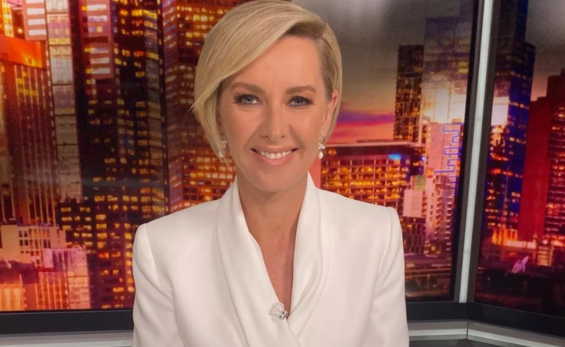 A person with short blonde hair in a white suit, smiling in front of a cityscape backdrop.