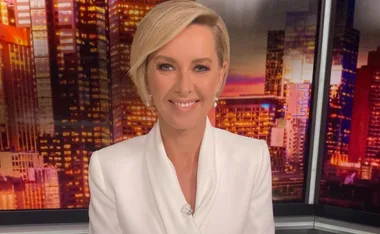 A person with short blonde hair in a white suit, smiling in front of a cityscape backdrop.