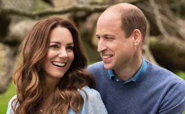A couple smiling at each other, standing outdoors with blurred trees in the background.