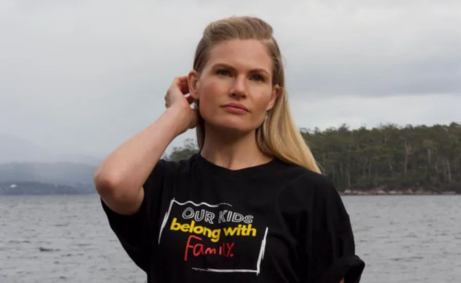 Woman wearing a "Our kids belong with family" T-shirt stands by a lake, touching her hair, with trees in the background.