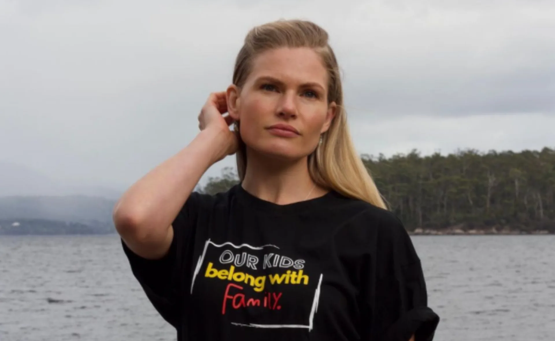 Woman wearing a "Our kids belong with family" T-shirt stands by a lake, touching her hair, with trees in the background.