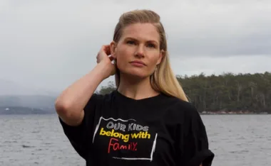 Woman wearing a "Our kids belong with family" T-shirt stands by a lake, touching her hair, with trees in the background.