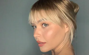 Blonde woman with bangs and blue eyes looking sideways against a gray background.