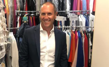 Man in blue suit smiling in a wardrobe filled with colorful clothes hanging on racks.