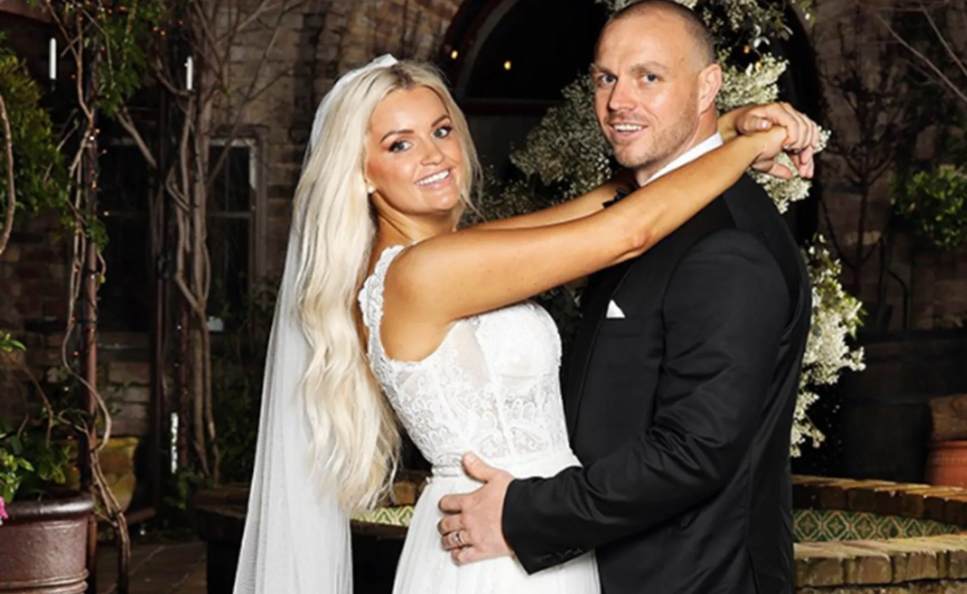Bride in white dress embraces groom in black suit, both smiling, standing in an outdoor setting with floral backdrop.