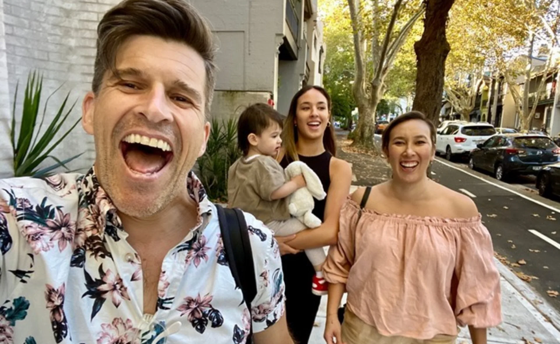 A man in a floral shirt and two women walk happily with a child holding a stuffed toy on a tree-lined street.