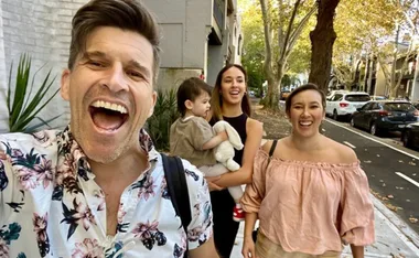 A man in a floral shirt and two women walk happily with a child holding a stuffed toy on a tree-lined street.