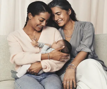 Two smiling women sit on a sofa, one holding a baby, signifying a joyful family moment.