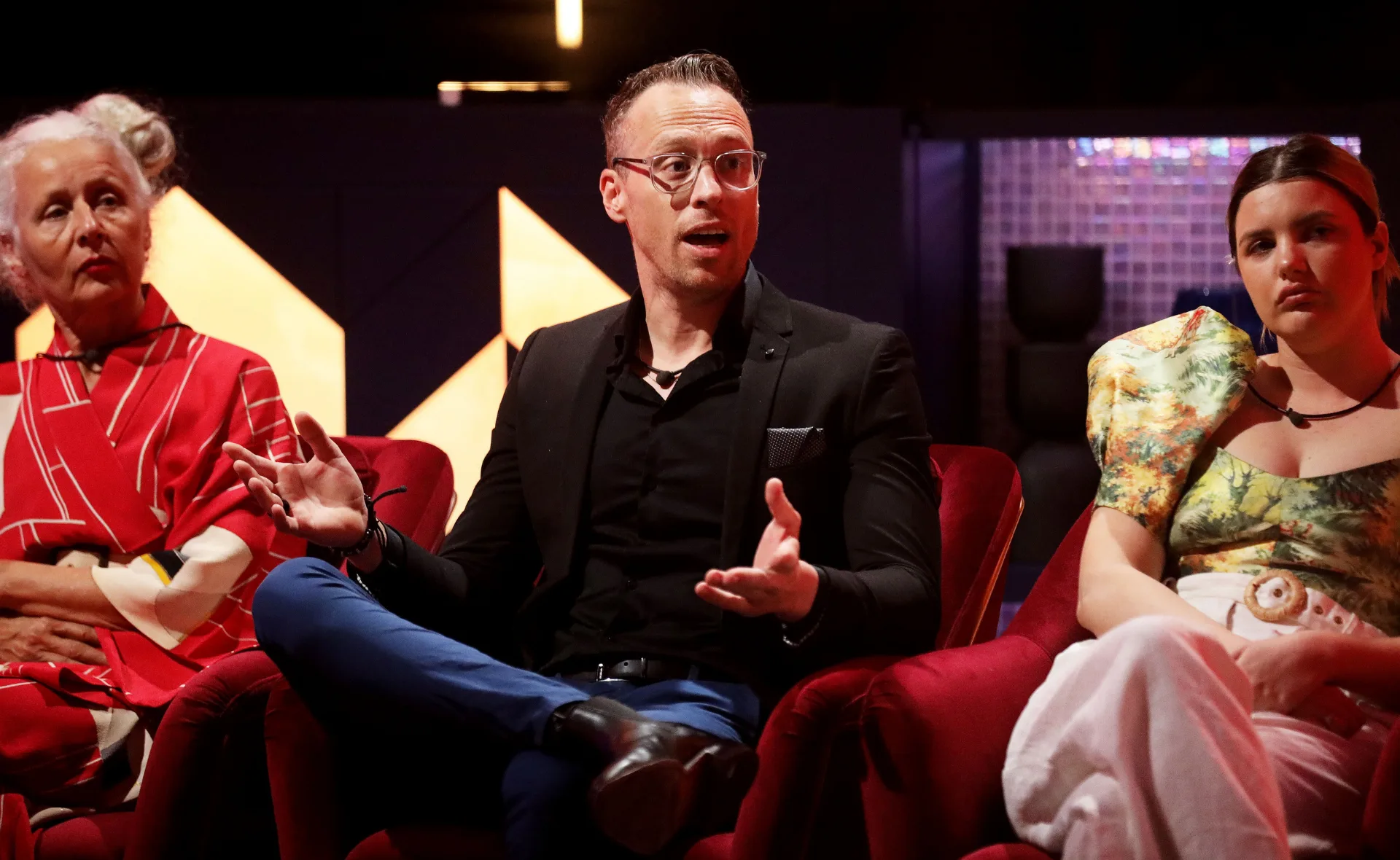 Three people sitting in red chairs talking, with a geometric background and warm lighting.