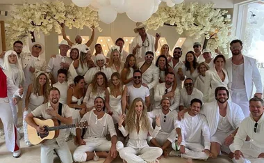 A group of people in all-white outfits pose together indoors, with decorative white flowers and balloons above them.