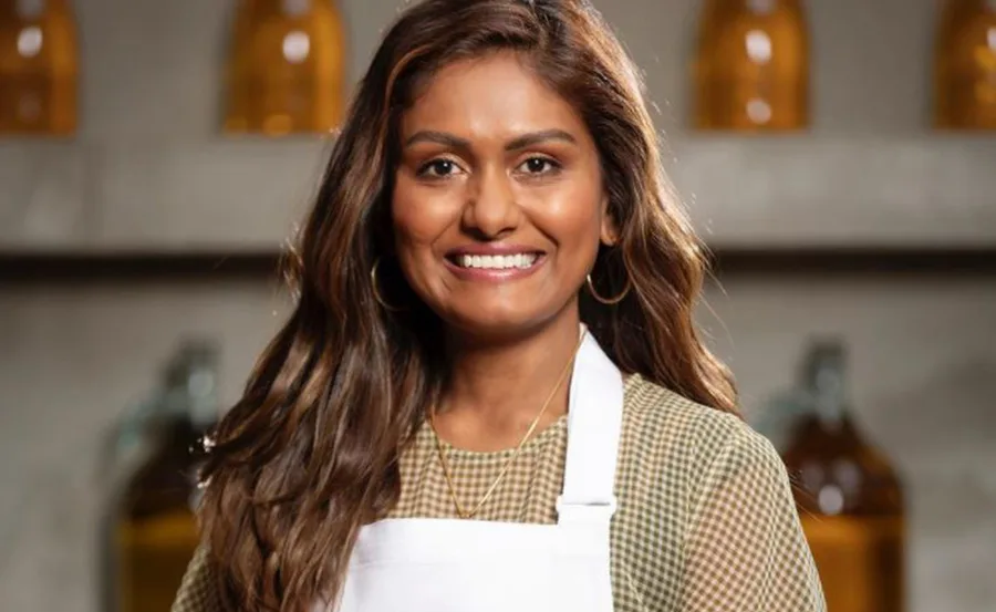 Person smiling, wearing a white apron, with long hair and hoop earrings, standing in a kitchen setting.