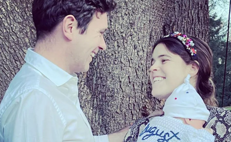 A couple smiling at their baby, who is wearing a knitted hat and wrapped in a blanket with "August" embroidered.