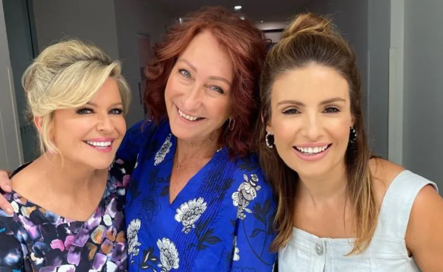 Three smiling women pose closely in a hallway, one with blonde hair, one with red hair, and one with brown hair.