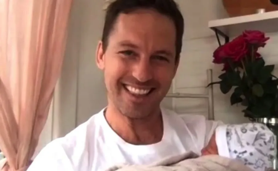 Man smiling while holding a baby wrapped in a blanket, with roses in the background.