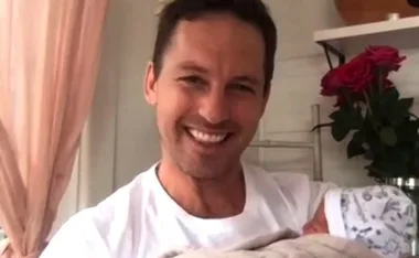 Man smiling while holding a baby wrapped in a blanket, with roses in the background.