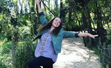 Woman joyfully posing on a forest path, wearing a green jacket and colorful shirt, surrounded by lush greenery.