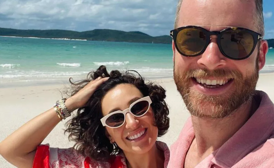 Couple taking a selfie on a sunny beach with turquoise water in the background, both wearing sunglasses and smiling.