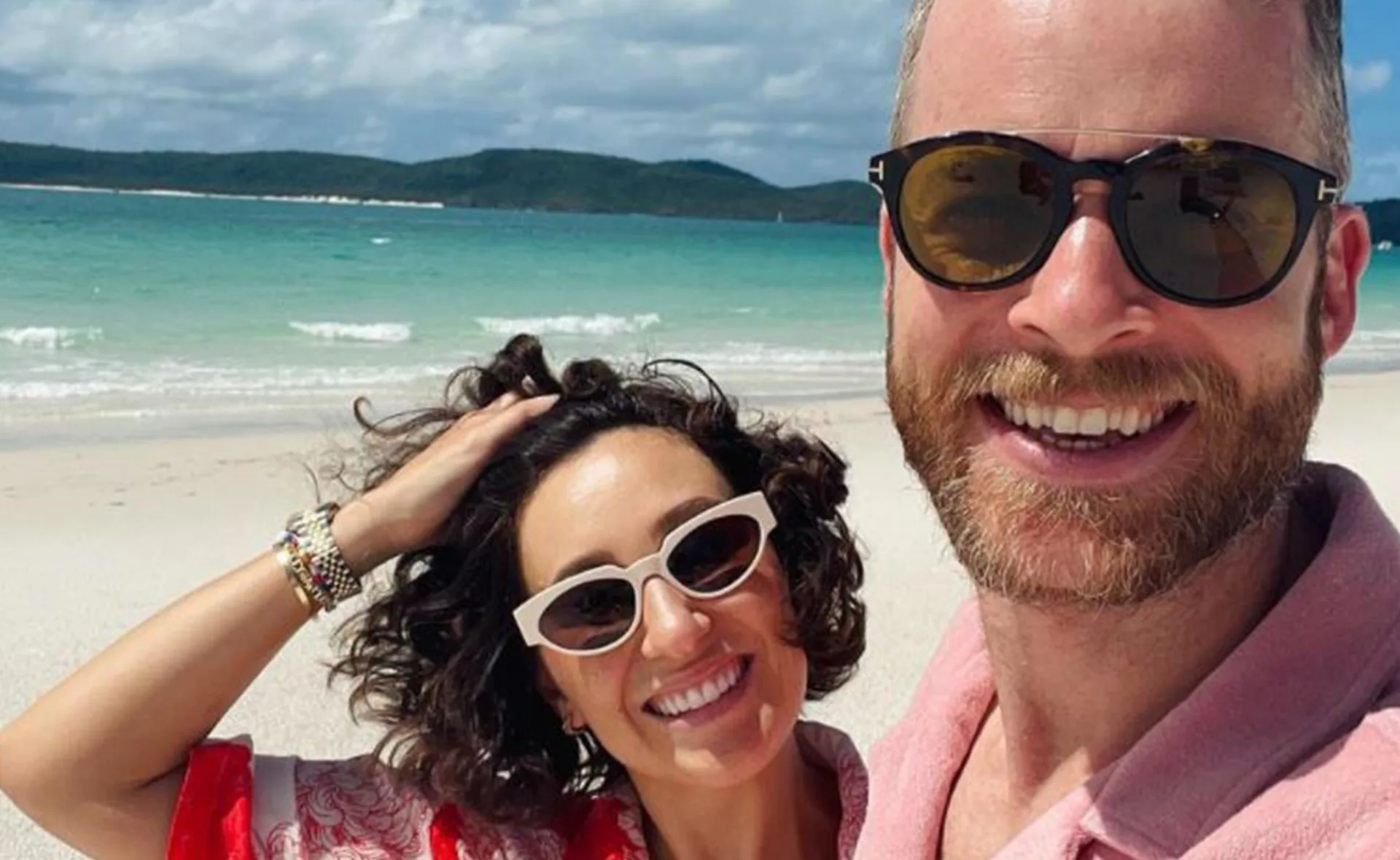 Couple taking a selfie on a sunny beach with turquoise water in the background, both wearing sunglasses and smiling.