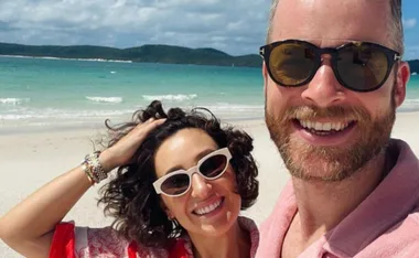 Couple taking a selfie on a sunny beach with turquoise water in the background, both wearing sunglasses and smiling.