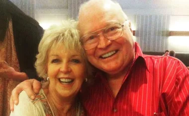 Elderly couple smiling and posing together, man in red striped shirt and woman with short blonde hair indoors.