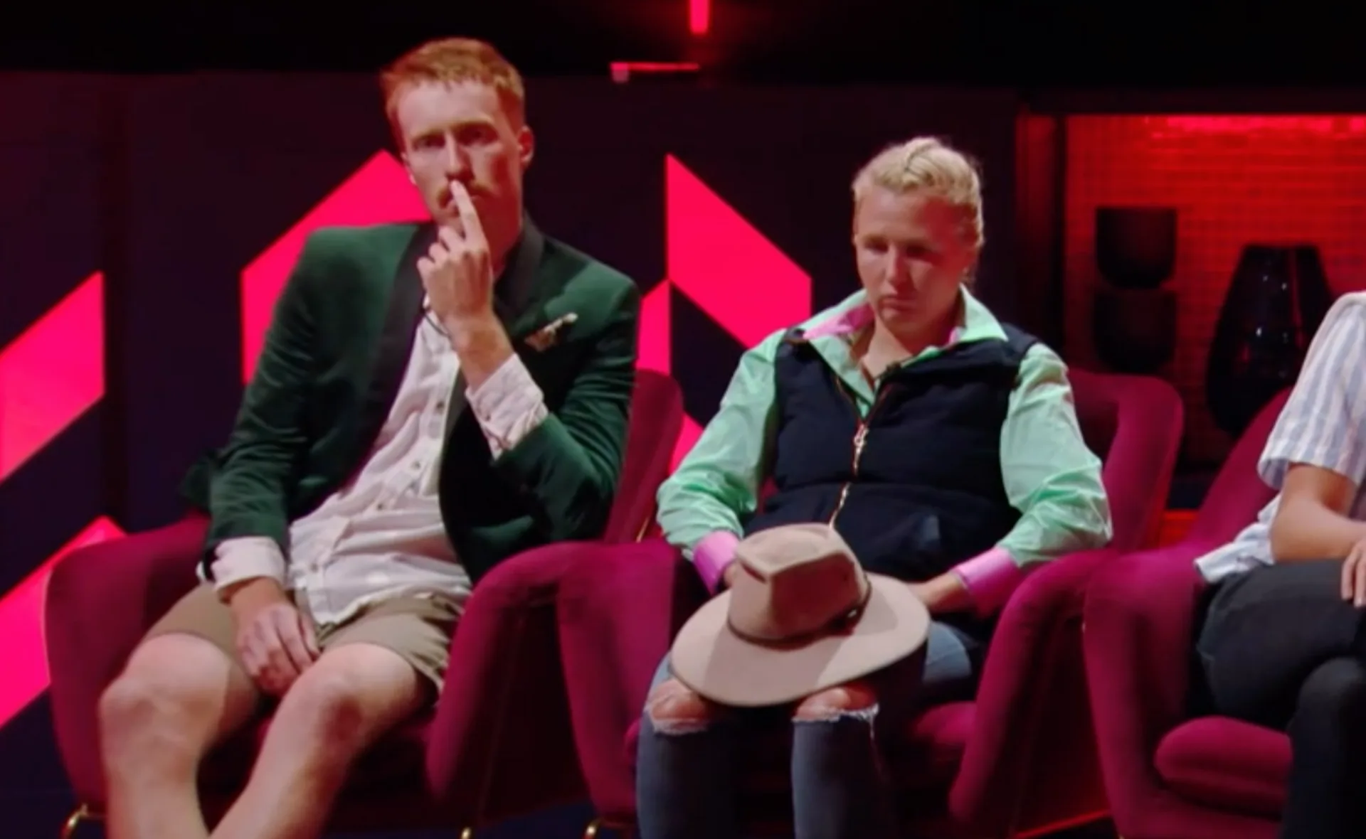 Two people seated on red chairs in a dimly lit room, one touching face, the other holds a hat, pink and red background.