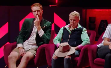 Two people seated on red chairs in a dimly lit room, one touching face, the other holds a hat, pink and red background.
