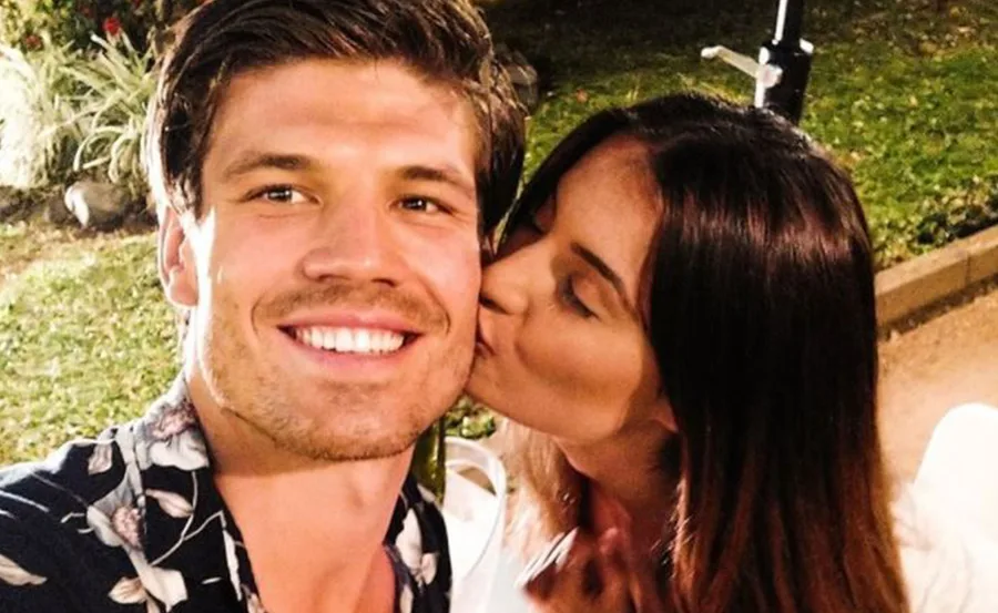 Man smiling with a floral shirt while a woman kisses his cheek outdoors.