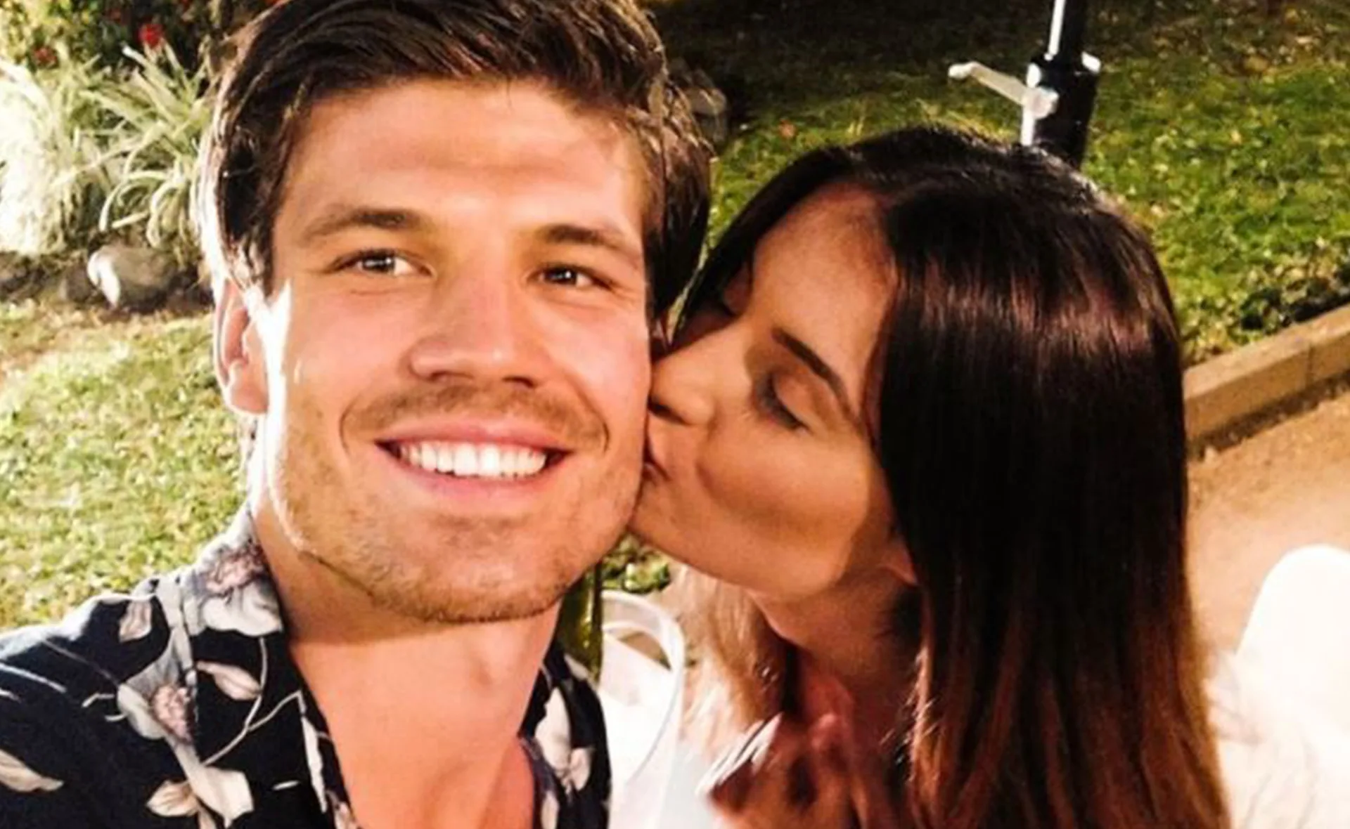 Man smiling with a floral shirt while a woman kisses his cheek outdoors.