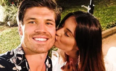 Man smiling with a floral shirt while a woman kisses his cheek outdoors.